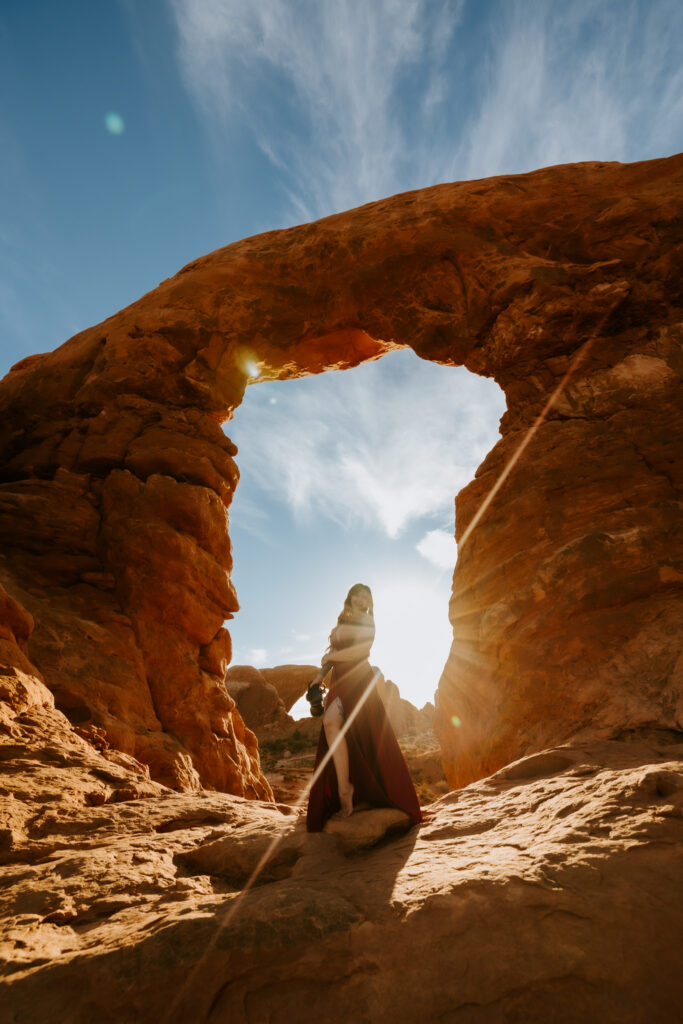 Bride in a flowing black wedding dress posing beneath a red rock arch with sunlight shining through behind her.