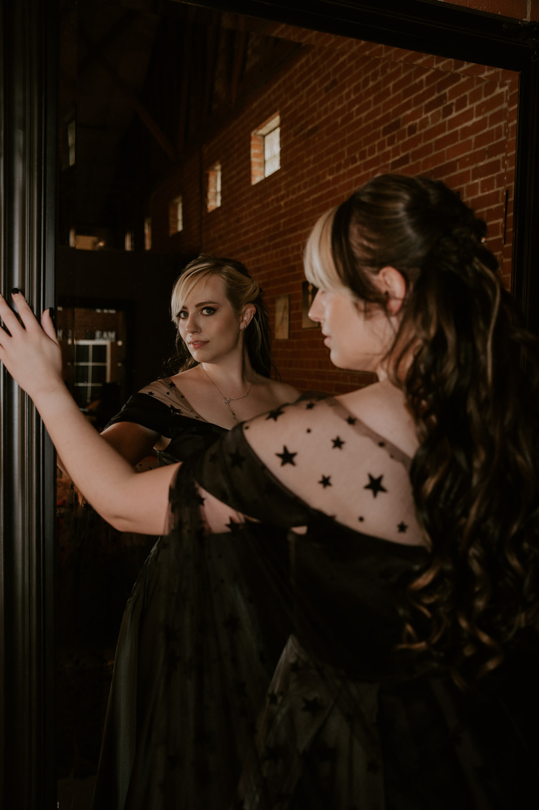 Bride in a black wedding dress with sheer star-covered sleeves looking at her reflection in a mirror.