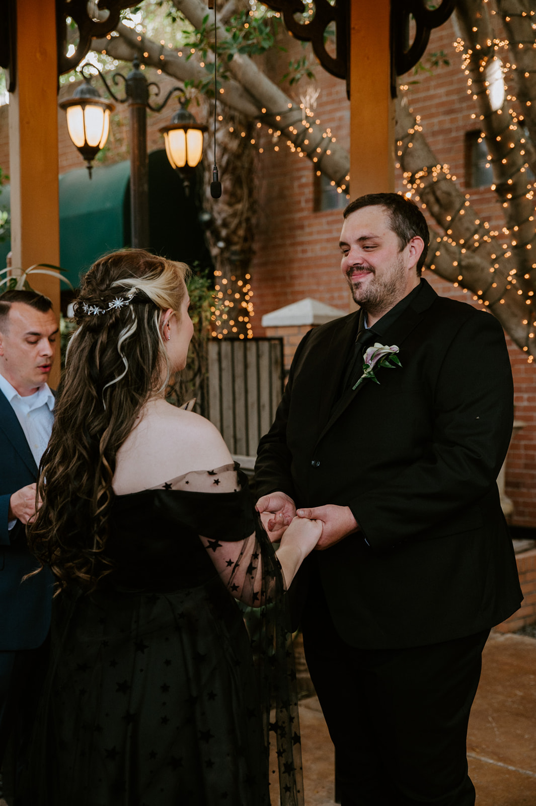 Couple holding hands during their non traditional wedding ceremony beneath string lights and trees.