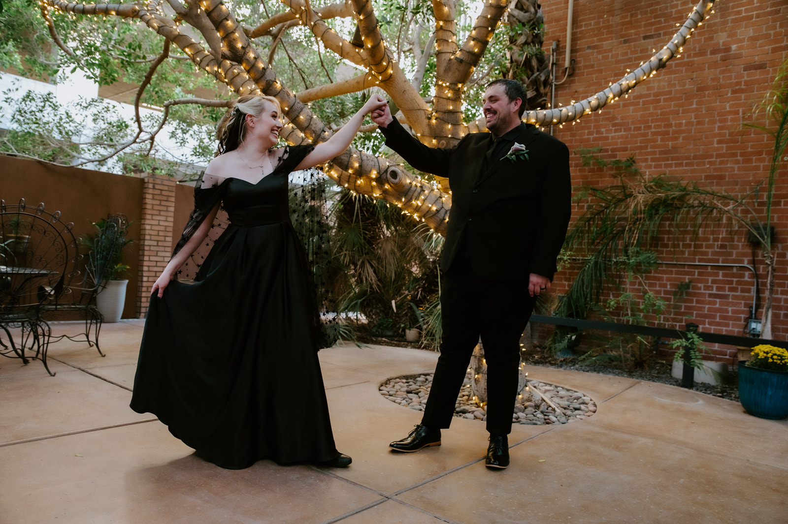 Bride in a black off-the-shoulder wedding dress twirling with her partner beneath a tree wrapped in string lights.