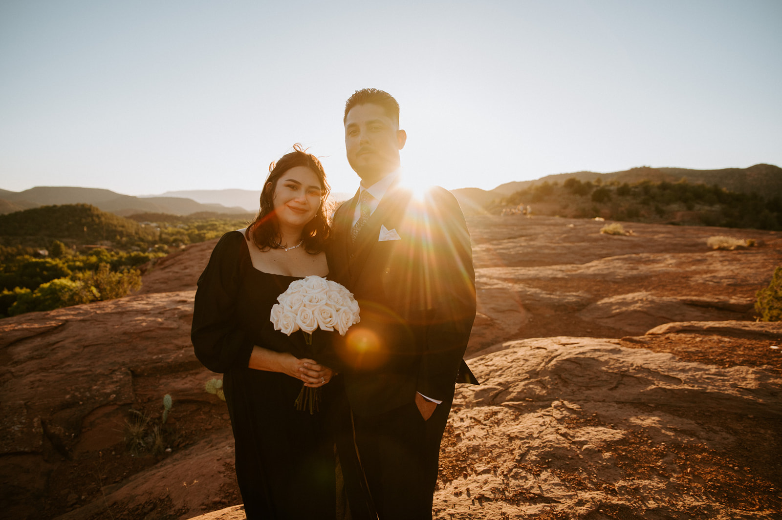 Newlyweds sitting together in a lush garden while the bride holds a white and purple rose bouquet with her black wedding dress.