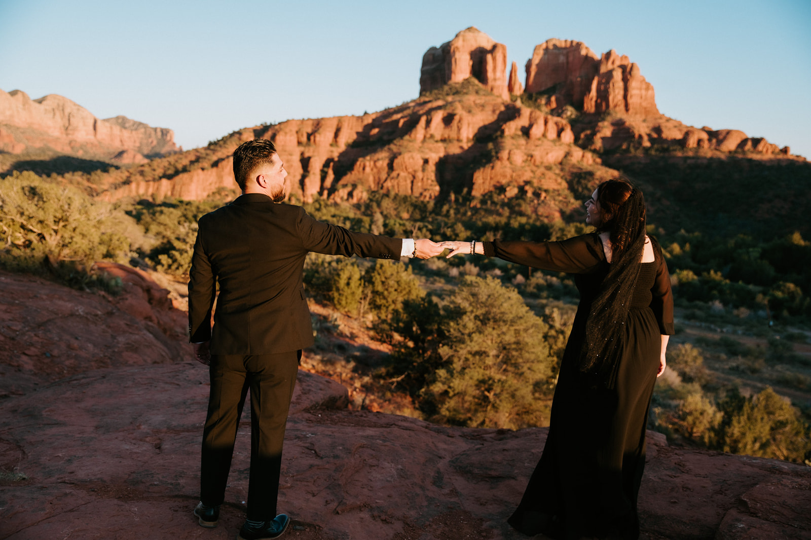 Couple holding hands across the red rocks during golden hour with dramatic Arizona mountains behind them.