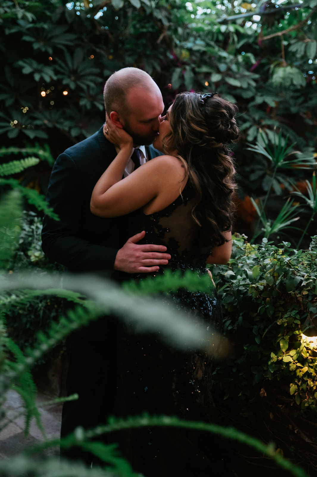 Newlyweds kissing among lush greenery while the bride wears a fitted black sequin wedding dress.
