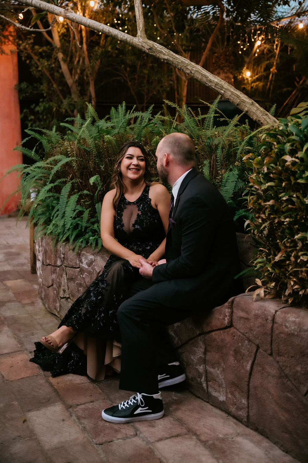 Newlyweds sitting together and laughing in a lush garden after their non traditional wedding, with the bride wearing a black sequin dress.