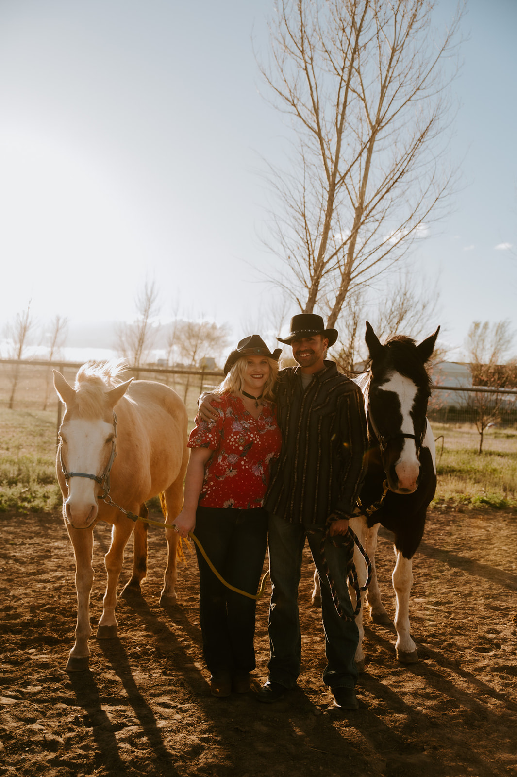 Couple wearing cowboy hats and posing with two horses in warm golden-hour light.