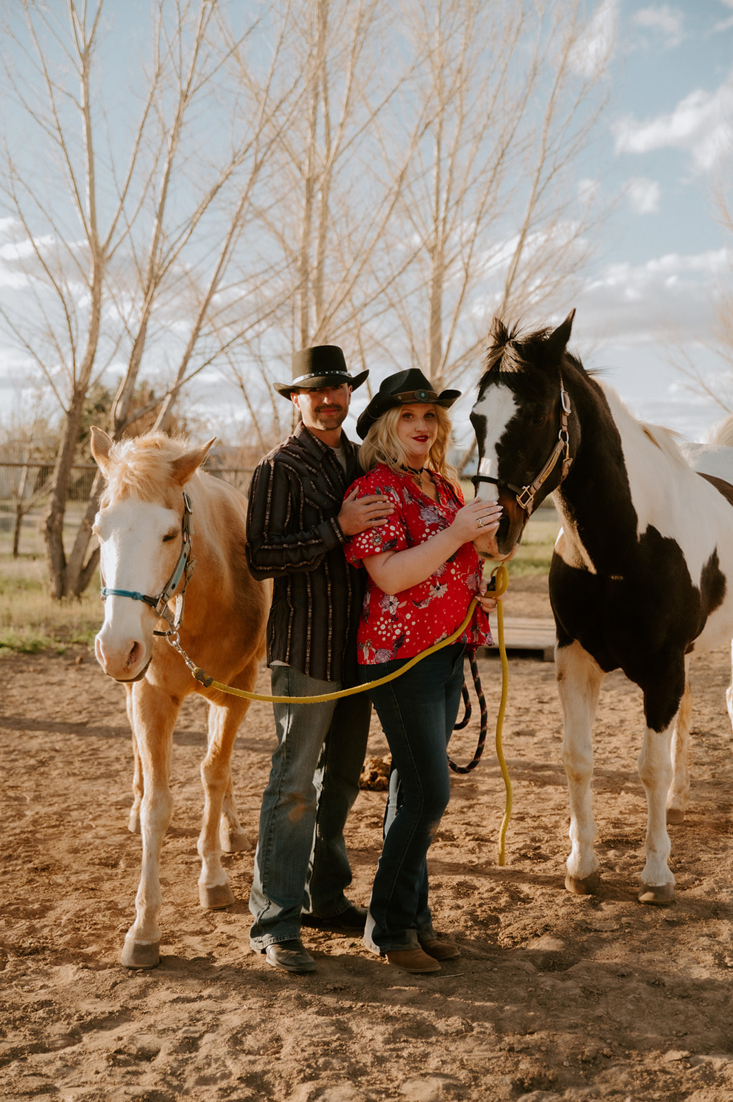 Couple posing between two horses in coordinated Western outfits, a playful example of what to wear for engagement photos.