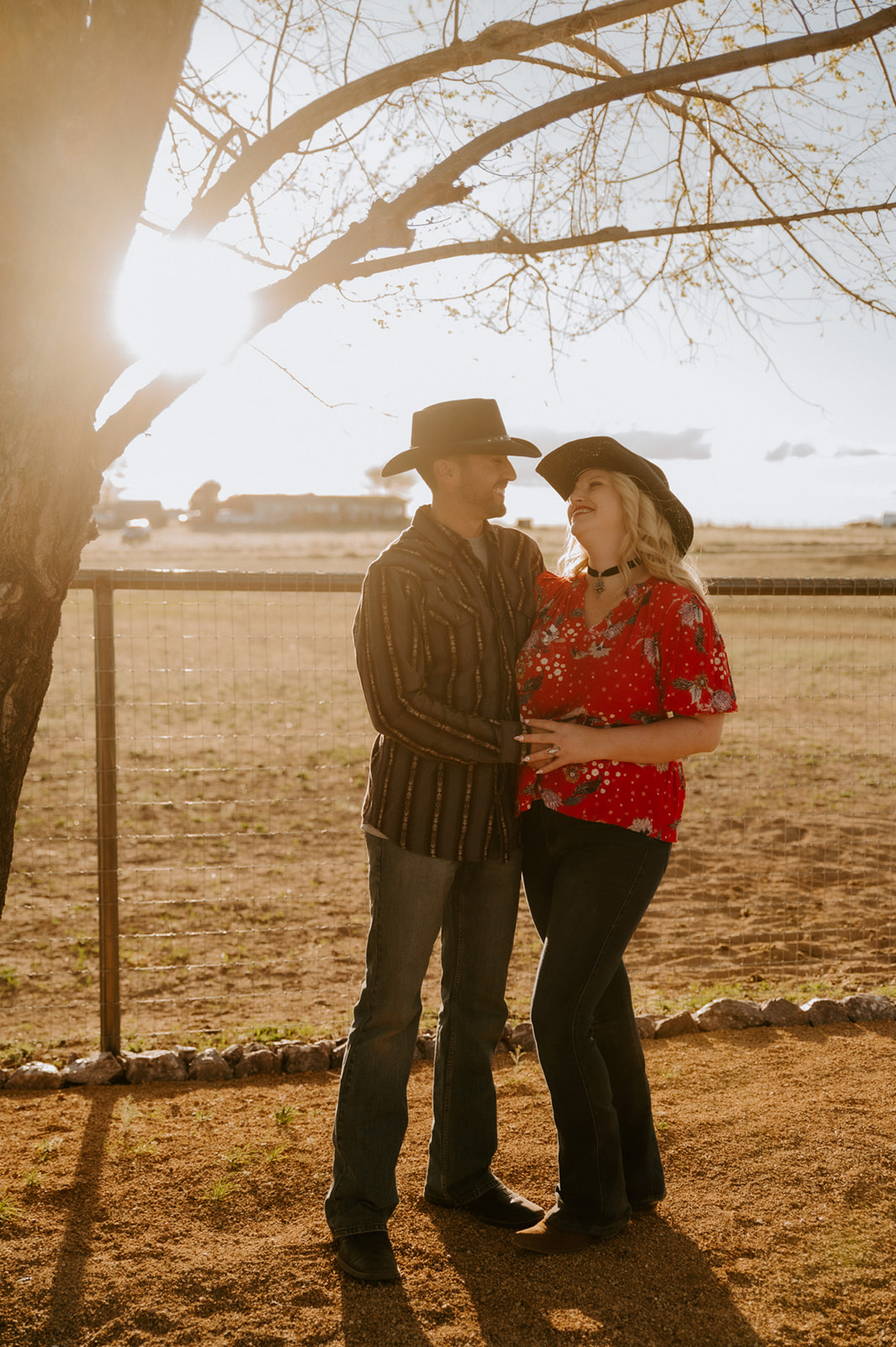 Couple wearing cowboy hats and Western outfits while smiling together beneath a tree at golden hour.