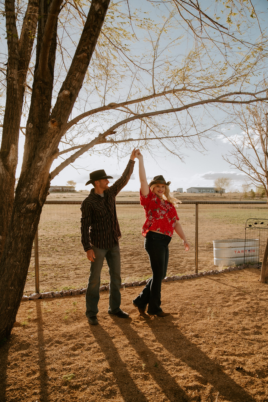 Couple dancing beneath a tree in cowboy hats and coordinated Western outfits, a fun example of what to wear for engagement photos.