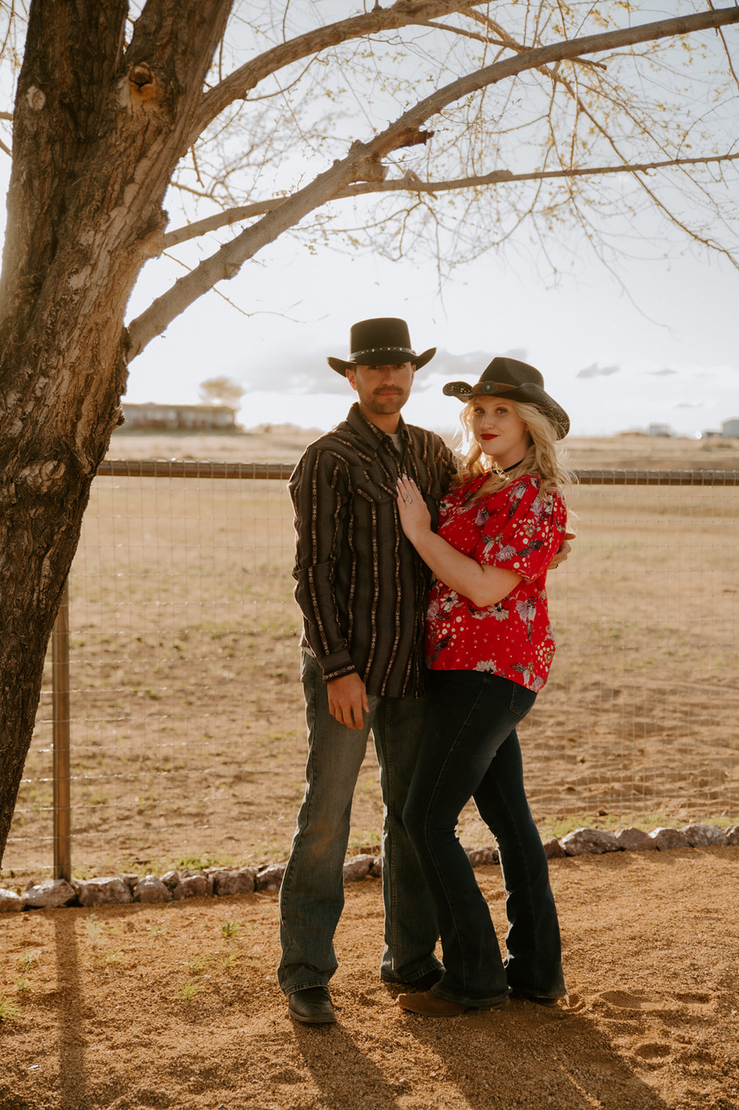 Couple posing beneath a large tree in coordinated Western outfits with cowboy hats and casual denim.