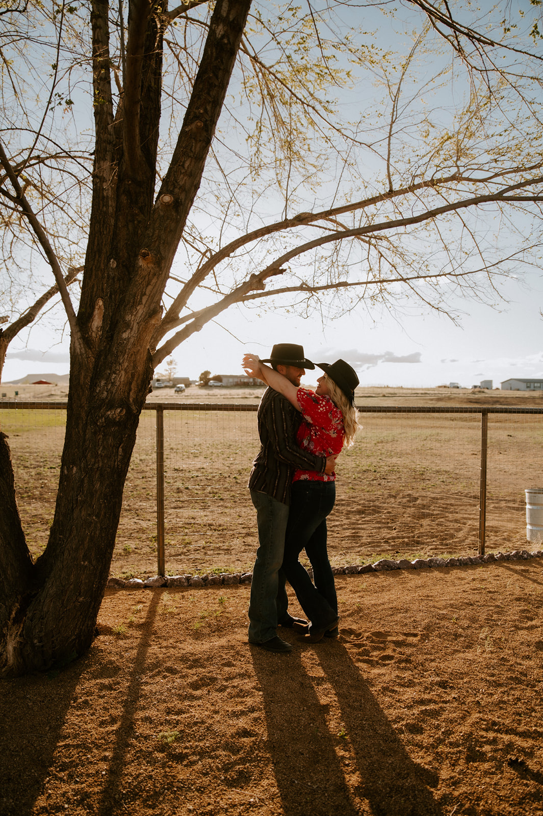 Couple embracing beneath a tree in cowboy hats and casual Western outfits, a playful example of what to wear for engagement photos.