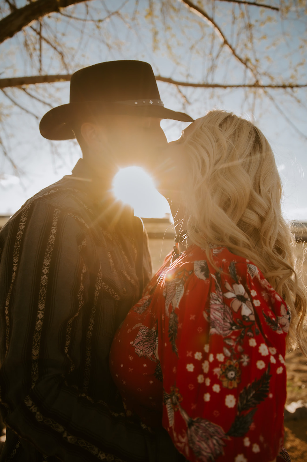 Couple kissing in warm golden sunlight while wearing cowboy hats and Western-inspired outfits.