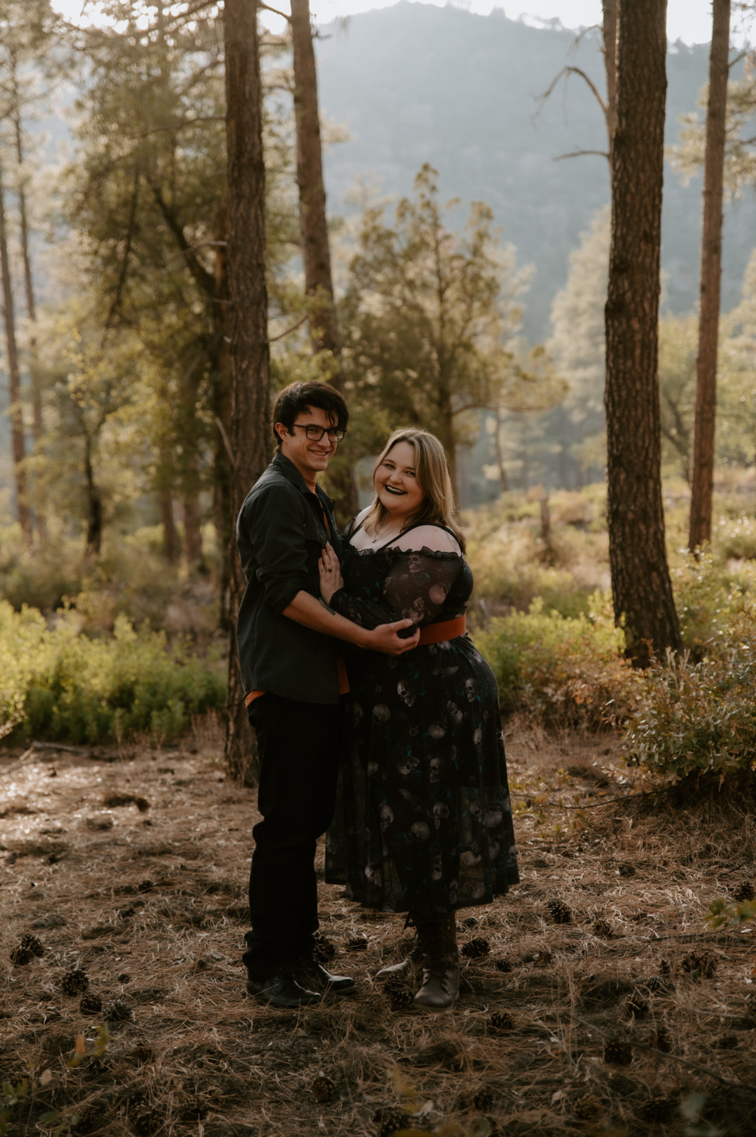 Smiling couple posing together among tall pine trees during their relaxed forest engagement session.