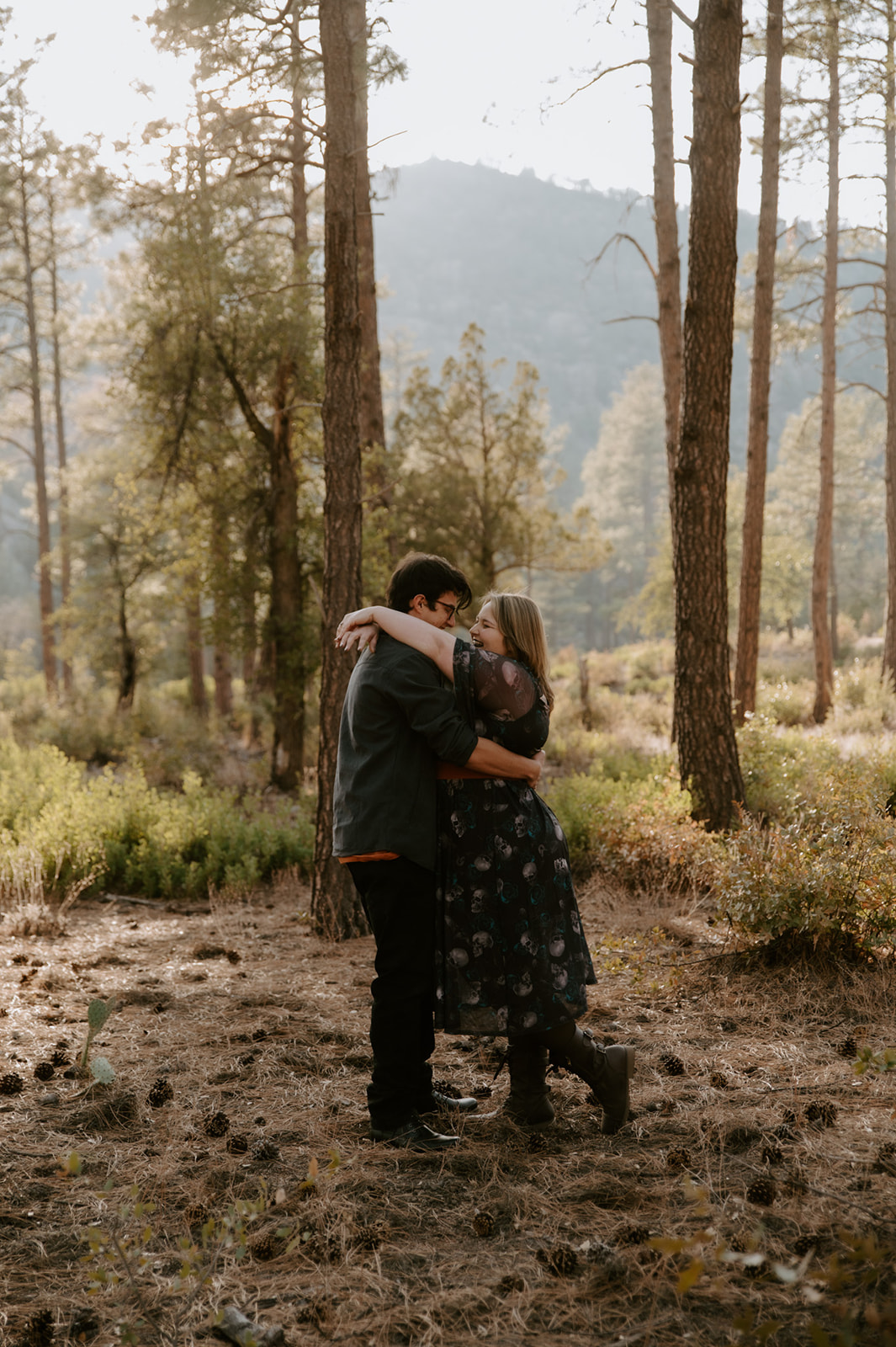 1. Couple embracing and laughing together among tall pine trees during their outdoor engagement session.