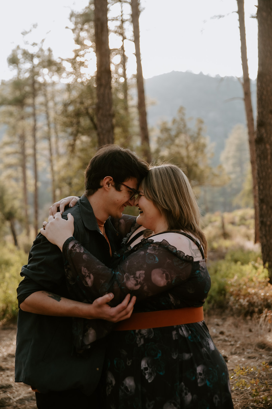 Couple embracing and smiling together among the pine trees during their outdoor engagement photos.