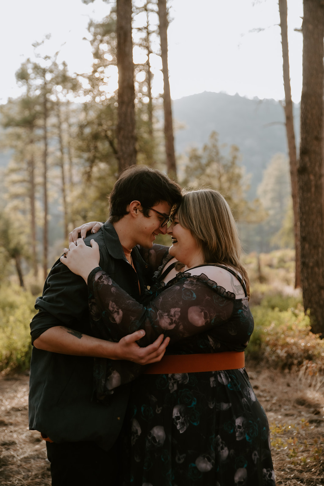 Couple laughing together in a forest, with a dark patterned dress and layered outfit inspiring what to wear for engagement photos.