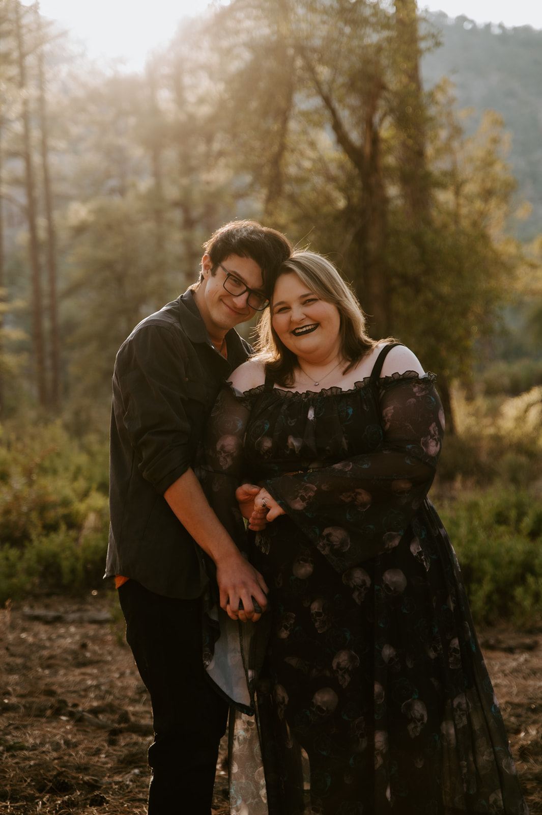 Engaged couple smiling in a pine forest, with a dark patterned dress and layered neutrals inspiring what to wear for engagement photos.
