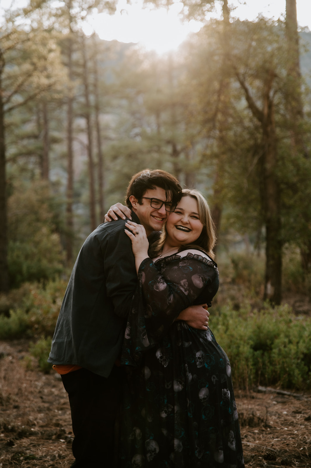 1. Smiling engaged couple embracing in a sunlit pine forest during their outdoor engagement session.