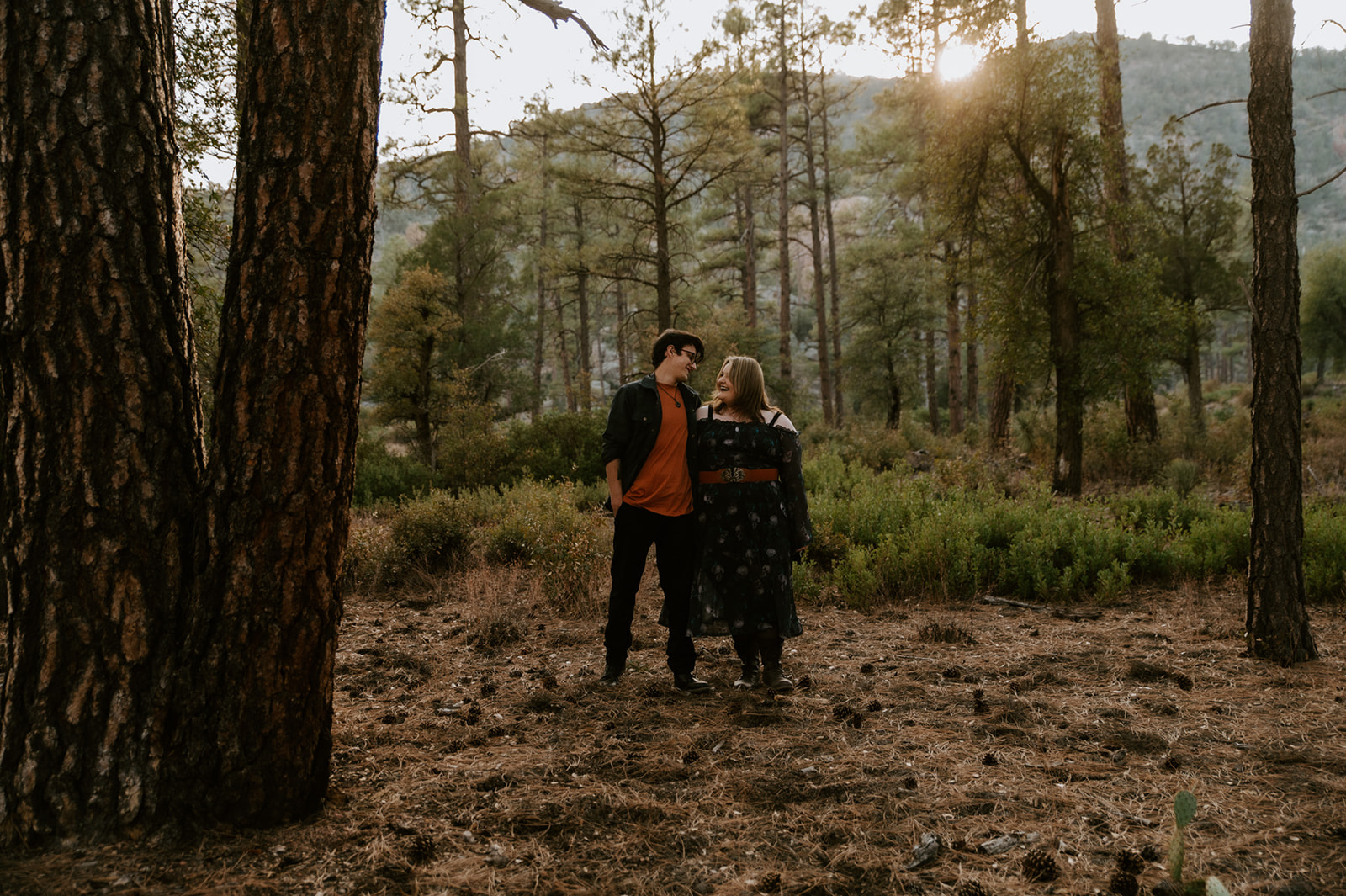 Couple standing together in a quiet pine forest with mountains and warm sunlight in the background.