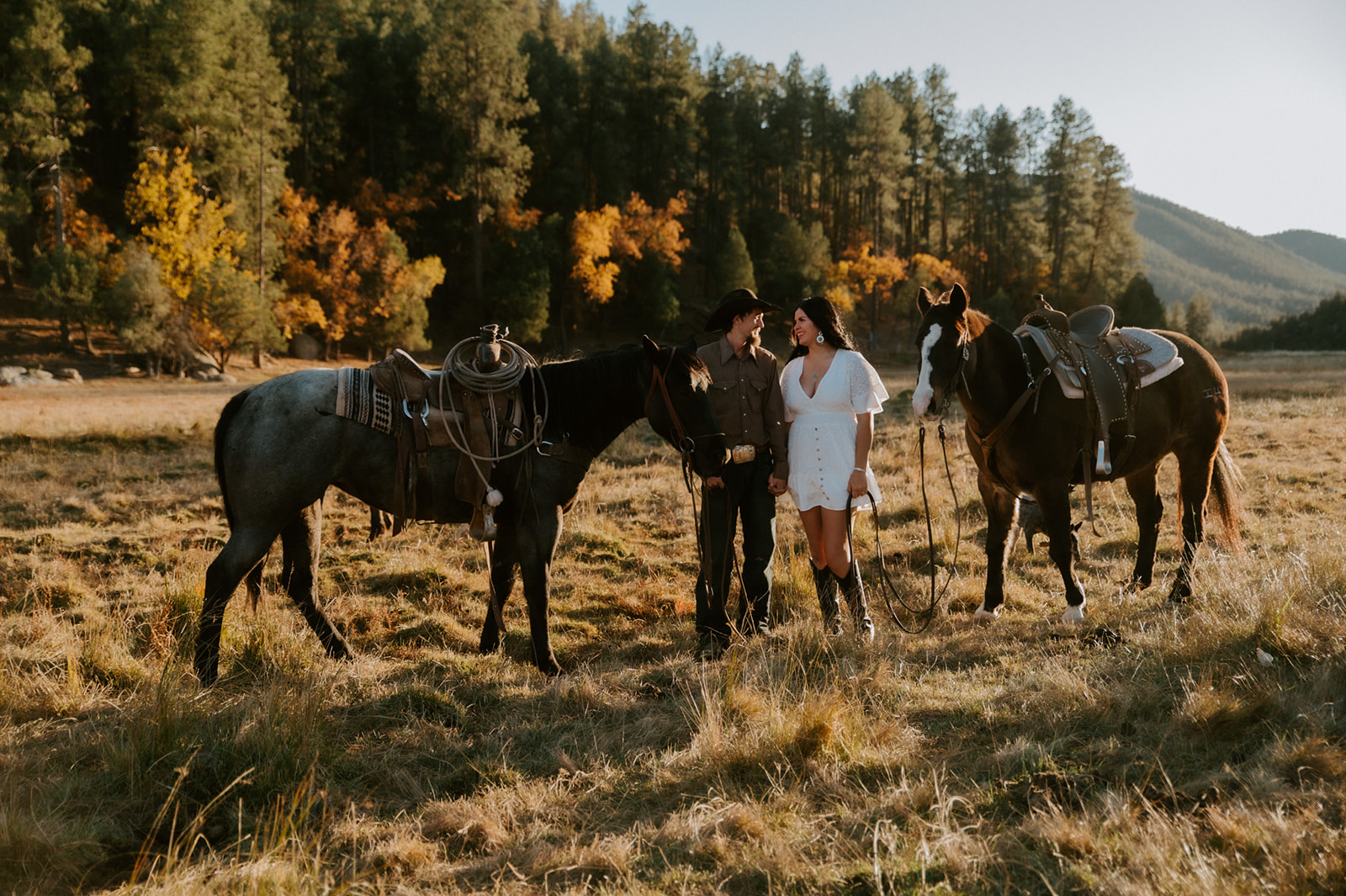 Couple posing with saddled horses in a golden meadow surrounded by autumn trees and mountains.