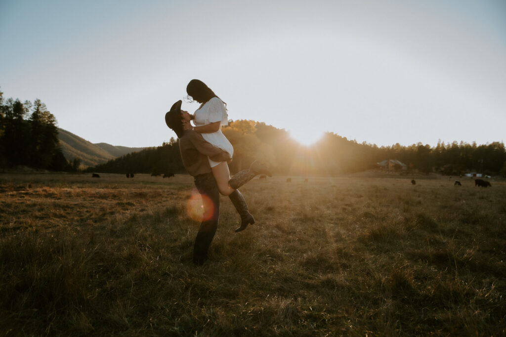 Man lifting his partner in a golden meadow, with a white dress and cowboy boots offering inspiration for what to wear for engagement photos.