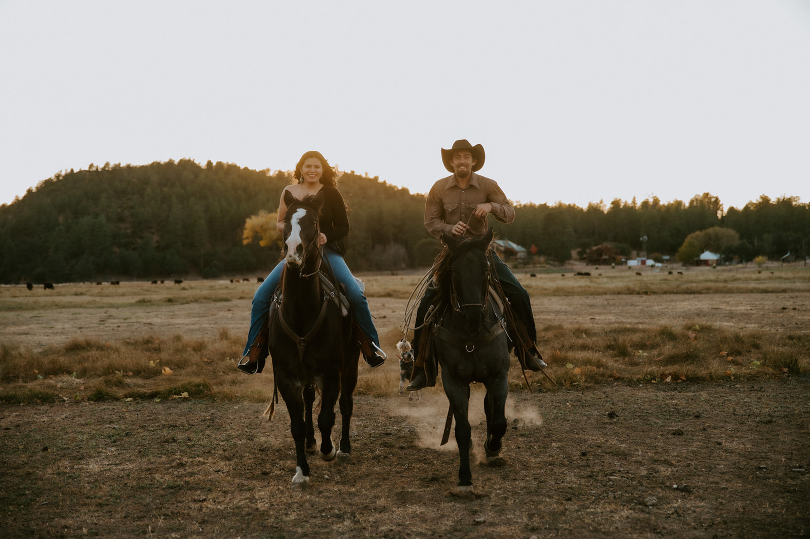 Engaged couple riding horses together across an open mountain meadow at sunset.