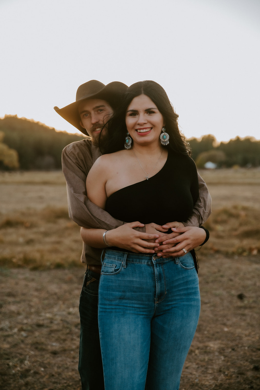 Couple embracing in an open field, showing how jeans, statement jewelry, and a cowboy hat can inspire what to wear for engagement photos.