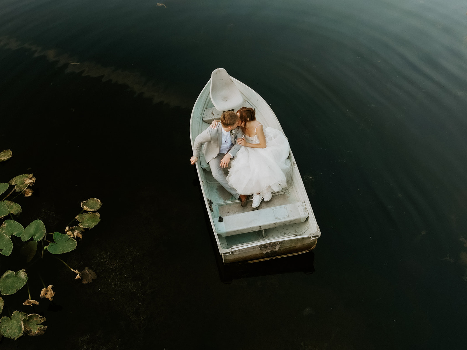 Aerial view of newlyweds cuddling in a small rowboat surrounded by dark, peaceful pond water.