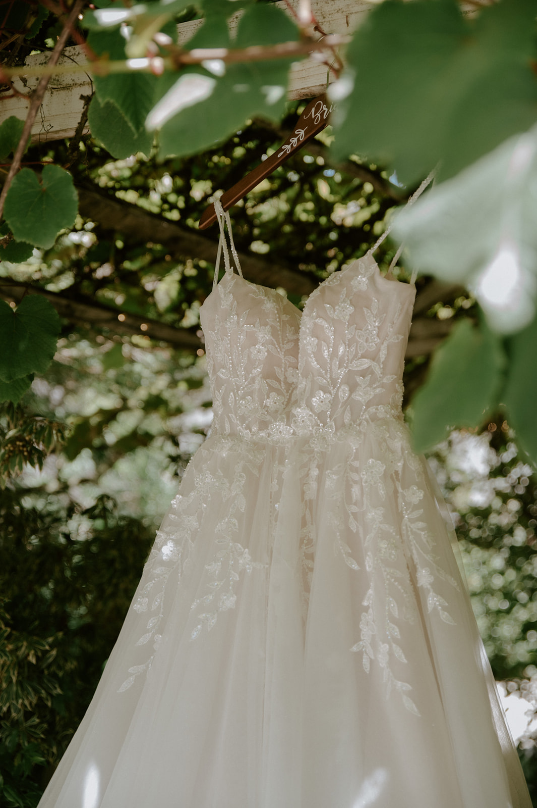Lace wedding dress with floral embroidery hanging from a wooden hanger beneath a leafy garden pergola.