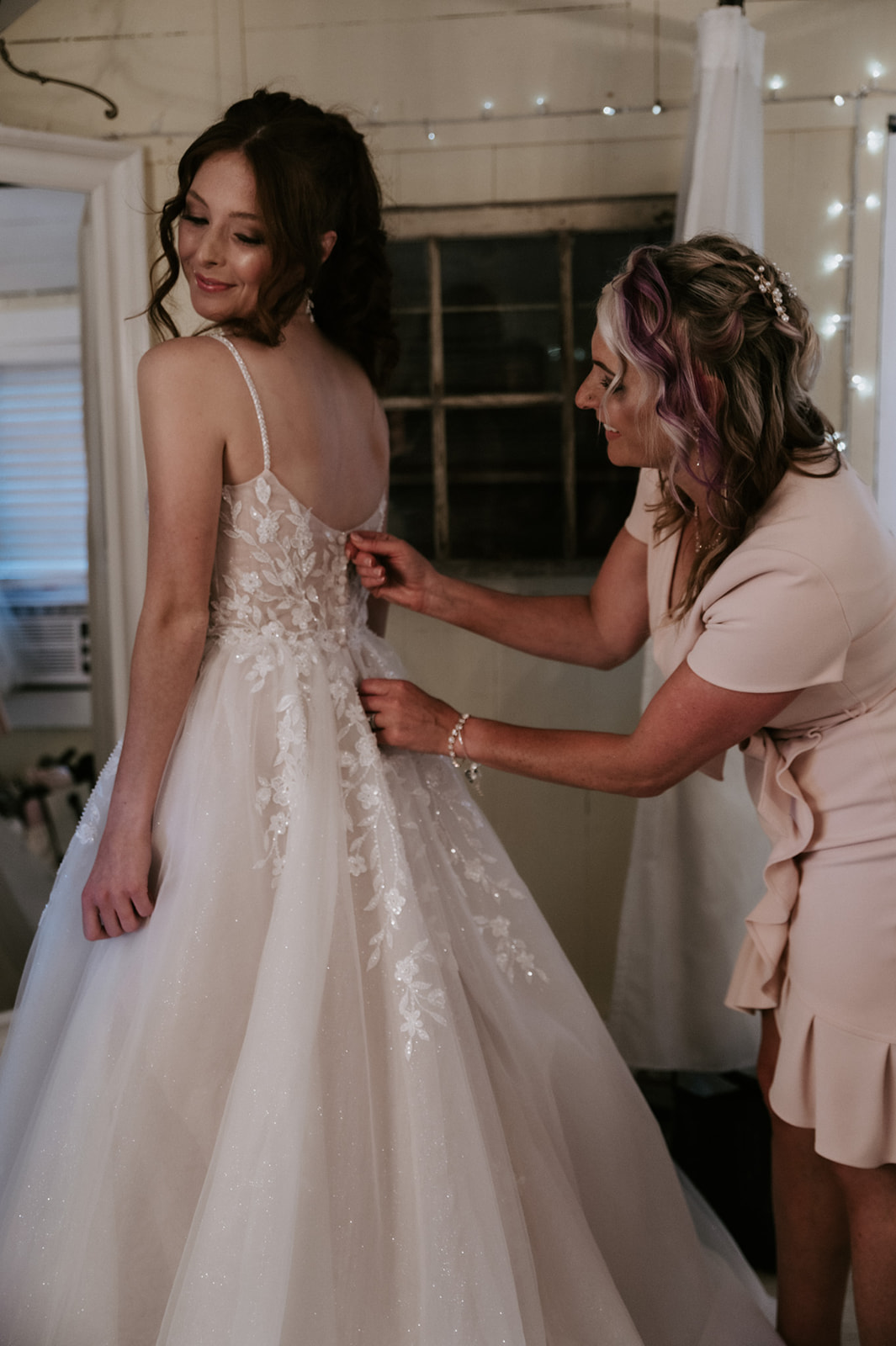Bride smiling as a woman helps fasten the back of her floral lace wedding dress before the ceremony.