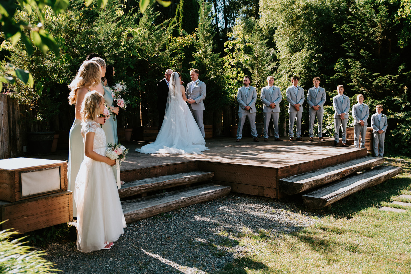 Outdoor wedding ceremony with the bride and groom exchanging vows on a wooden platform surrounded by bridesmaids and groomsmen.