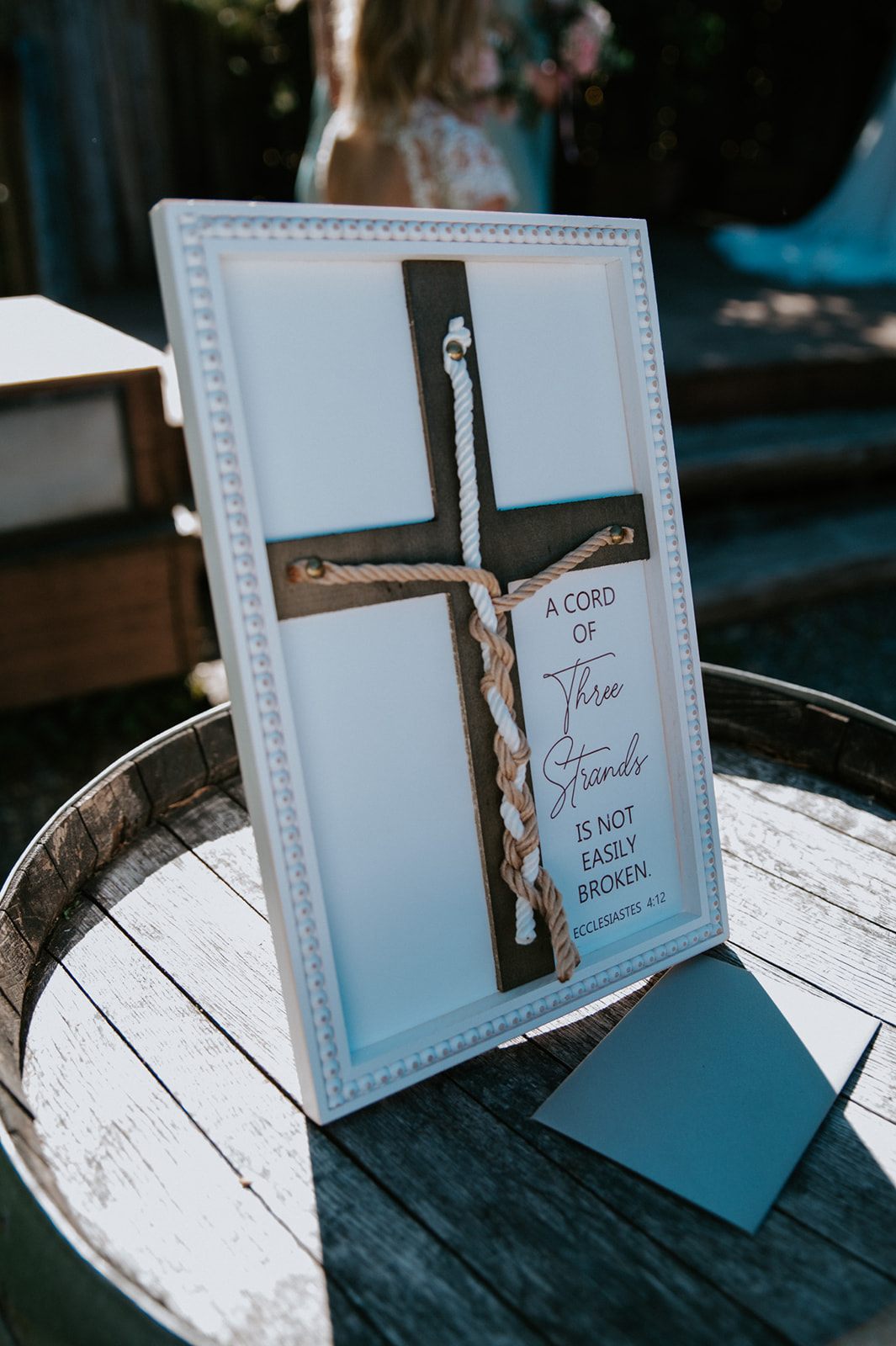 Framed wooden cross with a braided unity cord and Bible verse displayed on a rustic wine barrel during an outdoor wedding ceremony.