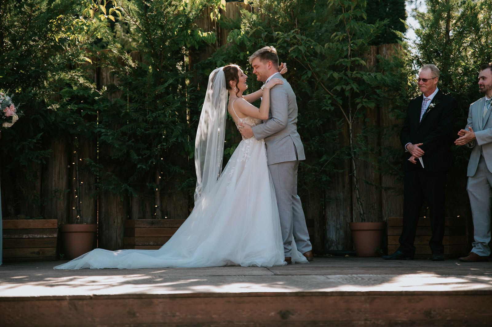 Bride and groom embracing on an outdoor ceremony platform surrounded by greenery and celebrating guests.