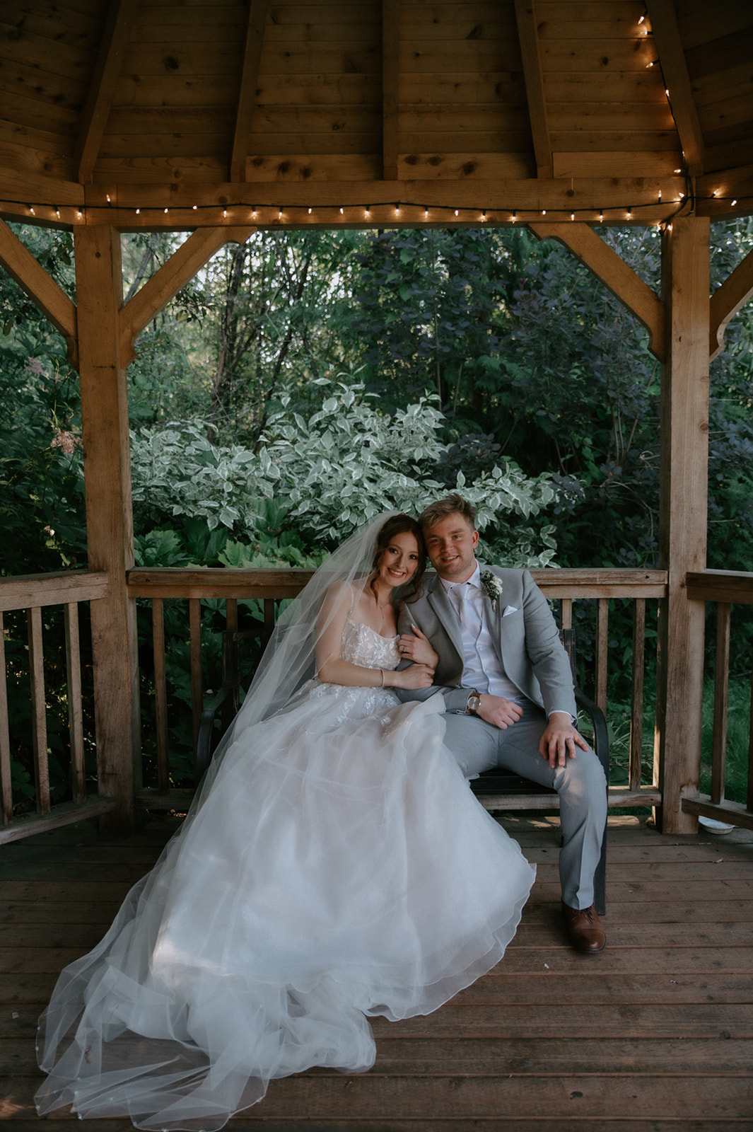 Newlyweds sitting together inside a wooden gazebo decorated with string lights and surrounded by lush greenery.