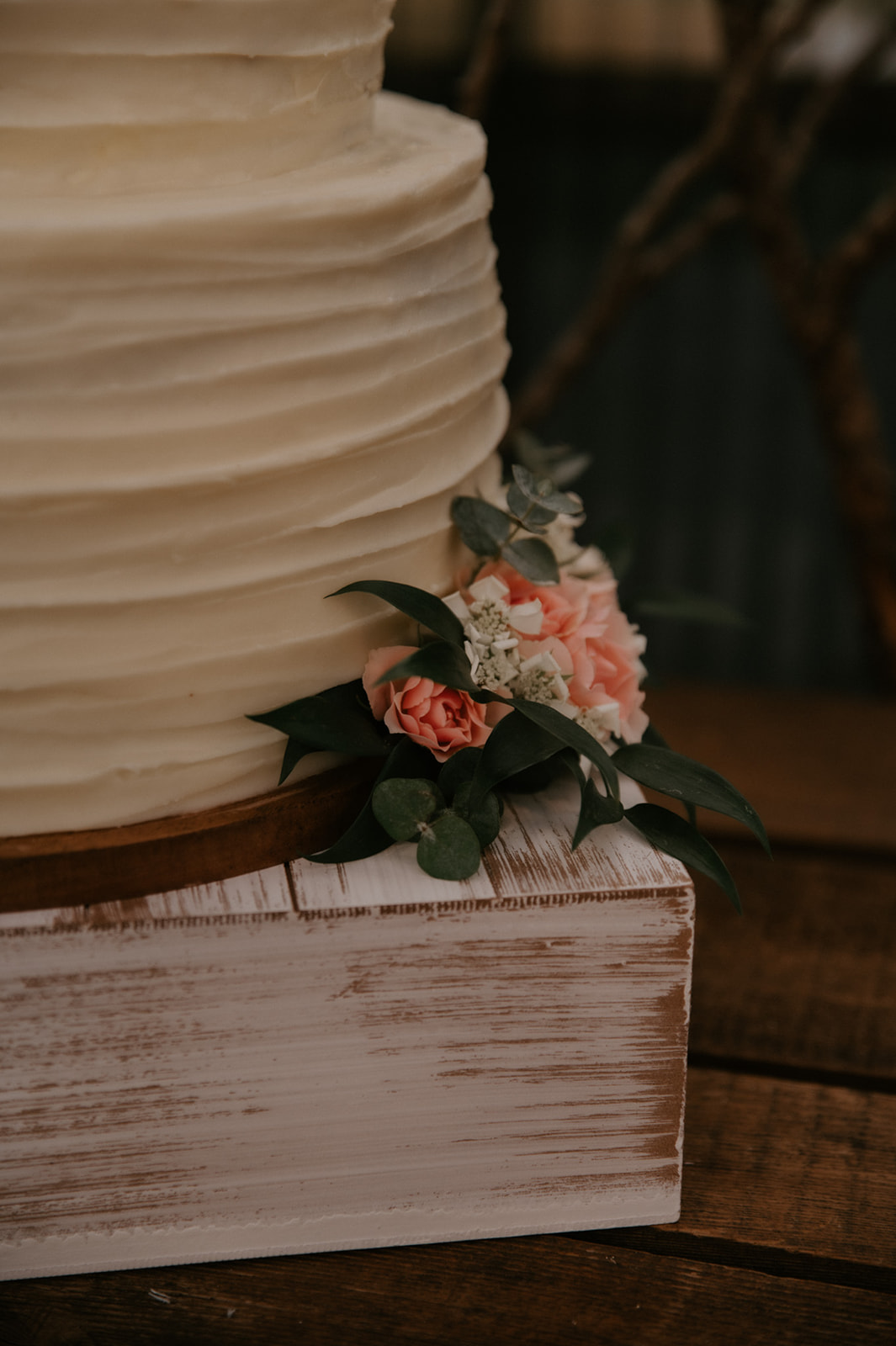 Close-up of a white tiered wedding cake decorated with blush pink roses and eucalyptus on a rustic wooden stand.