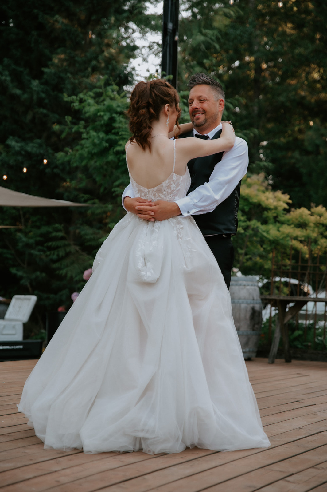 Bride in a flowing white gown dancing with a man in a black vest on an outdoor wooden dance floor.