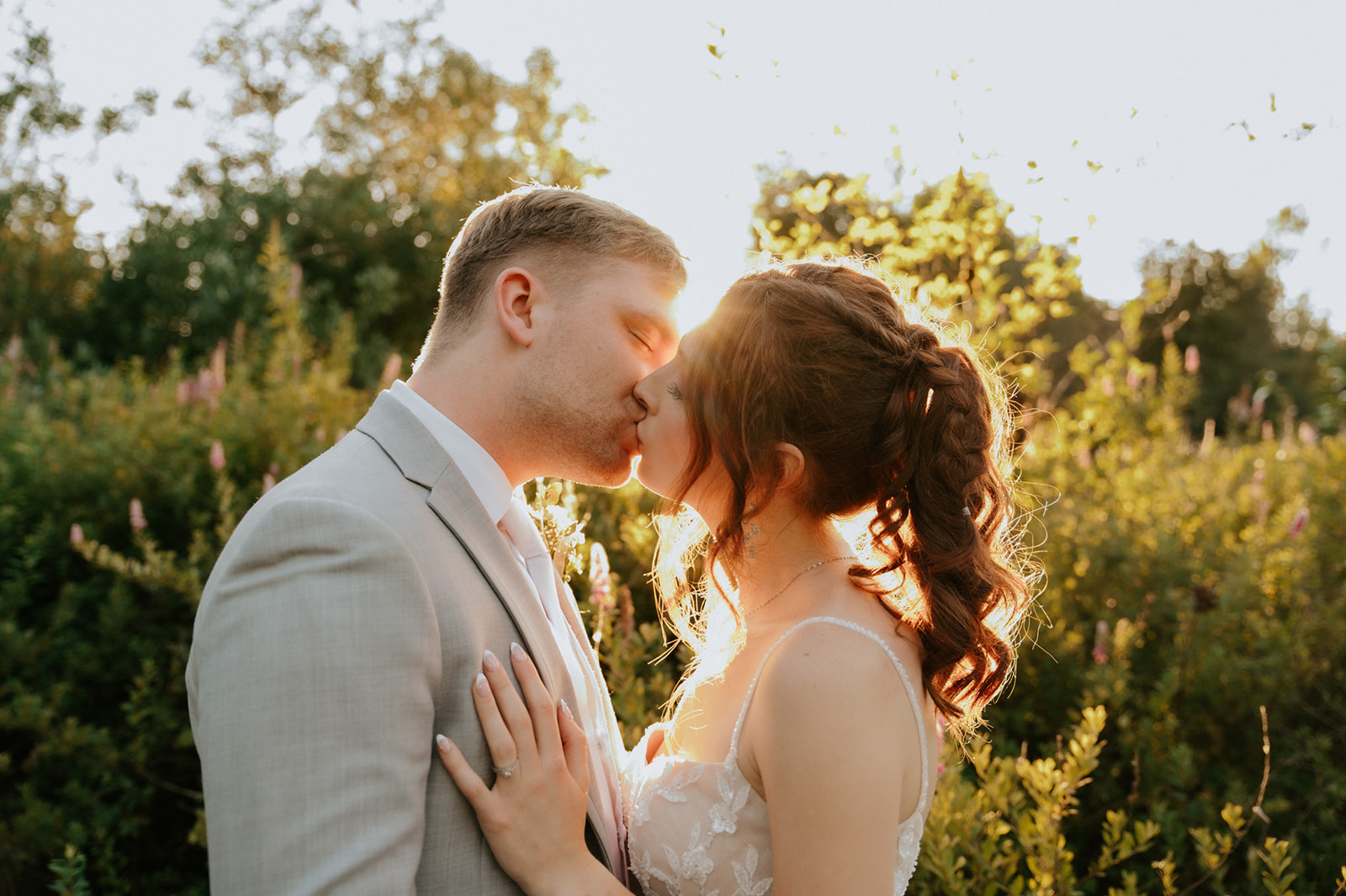 Newlyweds sharing a golden hour kiss surrounded by sunlit greenery and wildflowers.