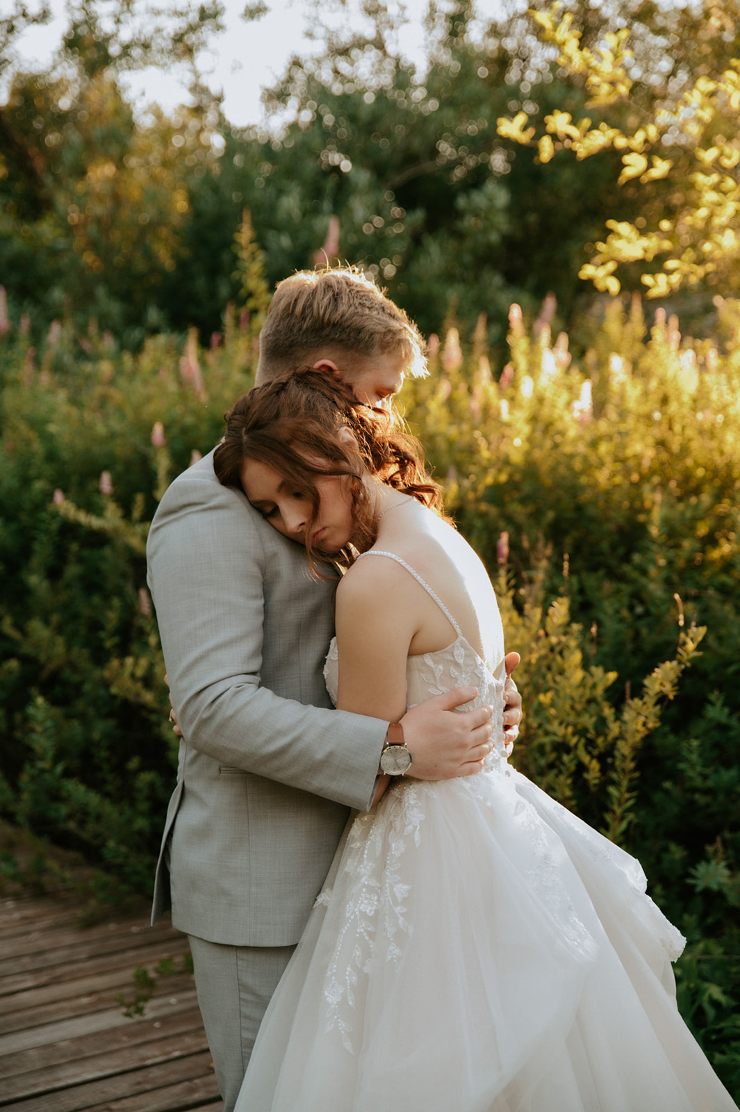 Bride resting her head against the groom's chest during intimate golden hour wedding portraits surrounded by greenery.
