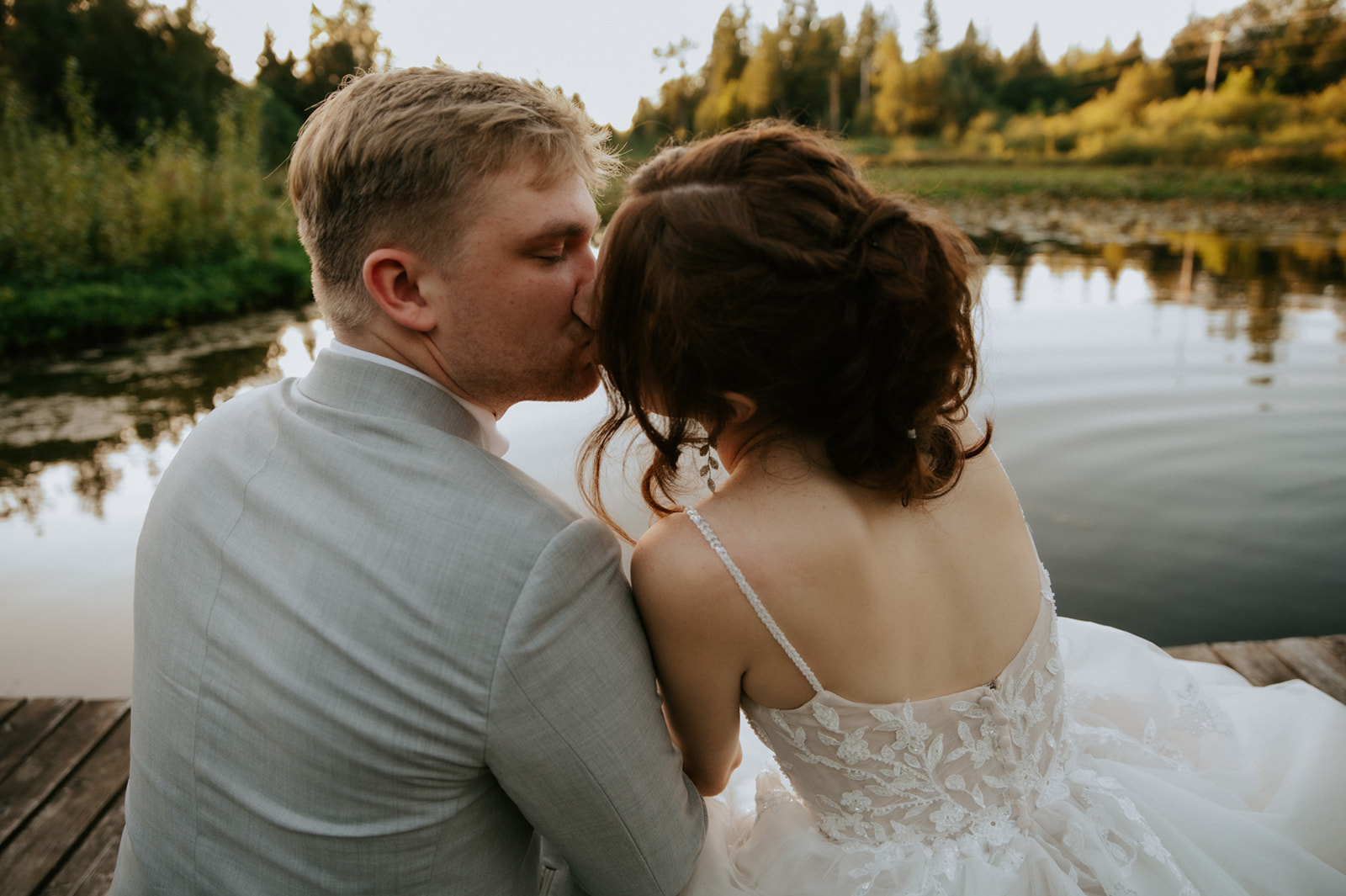 Bride and groom sharing a quiet kiss while sitting together on a wooden dock overlooking a peaceful pond.