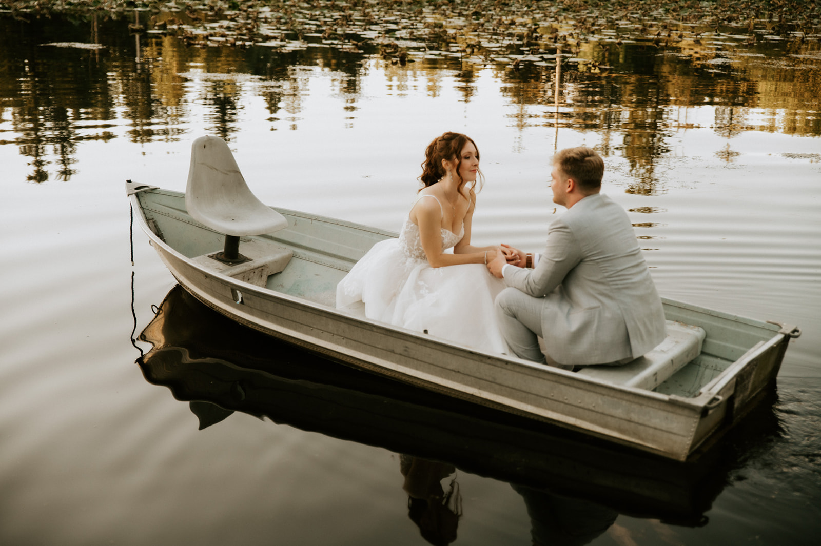 Bride and groom holding hands and smiling at each other while sitting together in a metal rowboat on calm water.