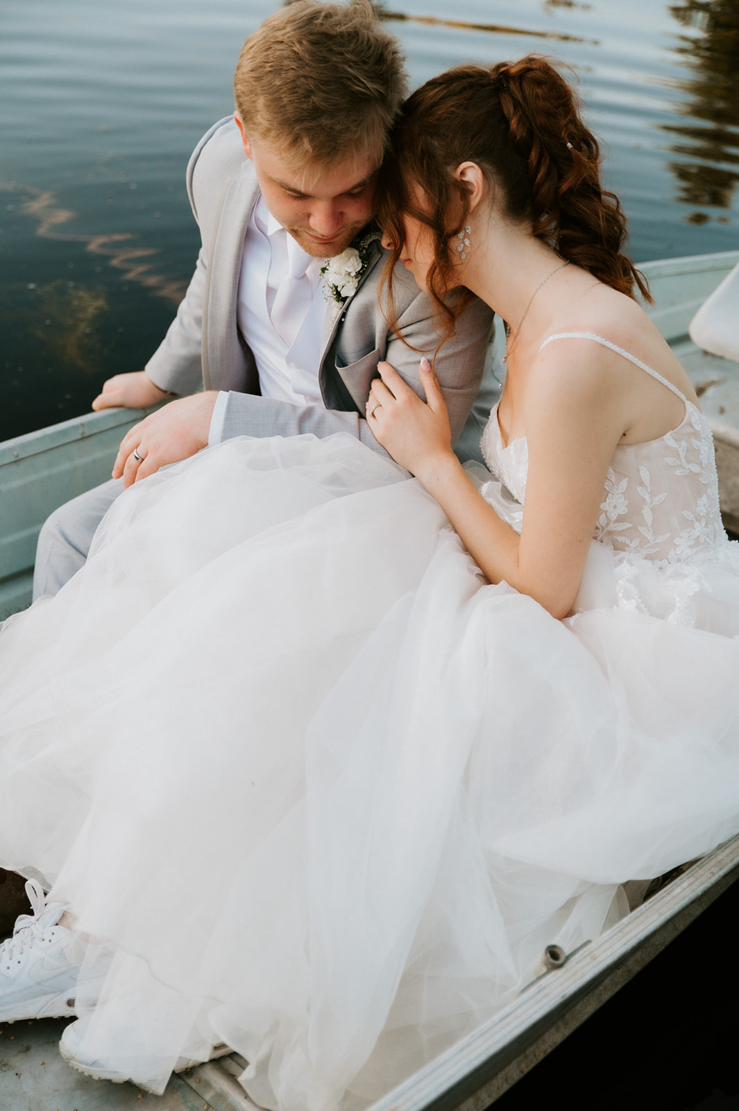 Close-up of newlyweds leaning their heads together in a rowboat, with the bride's flowing tulle gown draped across their laps.