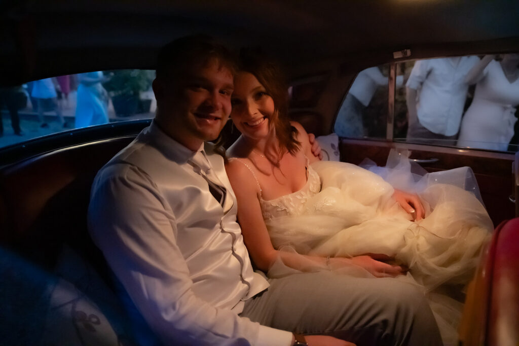 Smiling newlyweds sitting together in the back seat of a vintage car after their wedding celebration.